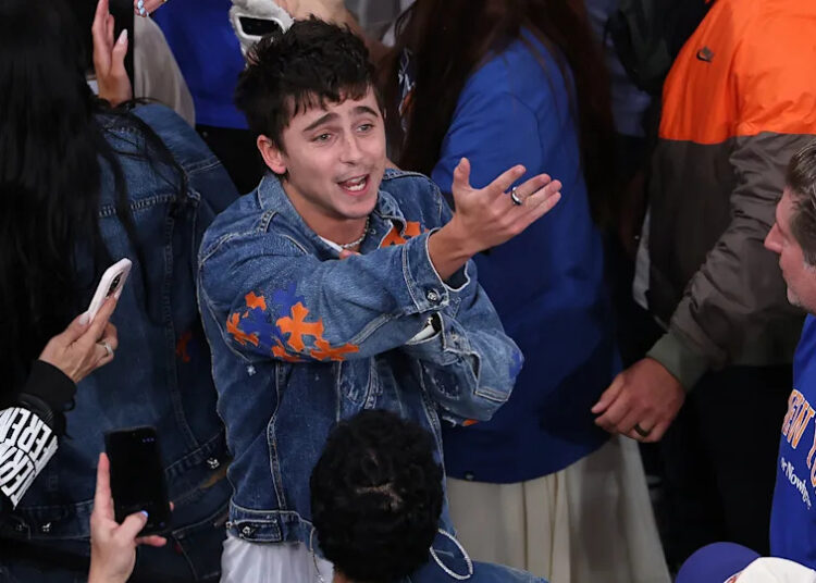 Timothée Chalamet in a denim jacket with orange accents gestures animatedly in a crowd at a game.