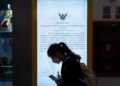 A woman wearing a mask walks past a video screen showing an official statement.
