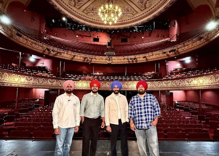 Rangle Sardar on the stage at the theatre. The four men stand in a line with the seats of the theatre behind them. They are all wearing shirts, jeans or black trousers and turbans.