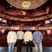 Rangle Sardar on the stage at the theatre. The four men stand in a line with the seats of the theatre behind them. They are all wearing shirts, jeans or black trousers and turbans.