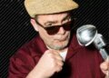 Trevor Carey, wearing a straw fedora and sunglasses, holding a vintage microphone and making a fist.