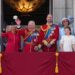 The British royal family in official dress stand on a balcony waving to a crowd.