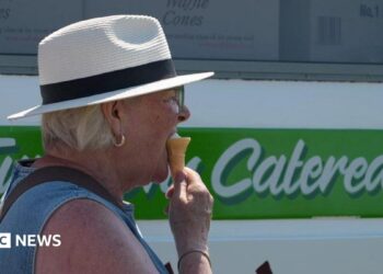 A woman with blonde hair standing next to an ice cream van. She is wearing a straw hat and a denim top. She is turned to the side facing towards the right and is eating an ice cream in a cone.