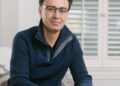 A man with glasses wearing a navy blue sweater sits on a couch, hands clasped, in a room with white shutters and a window in the background.