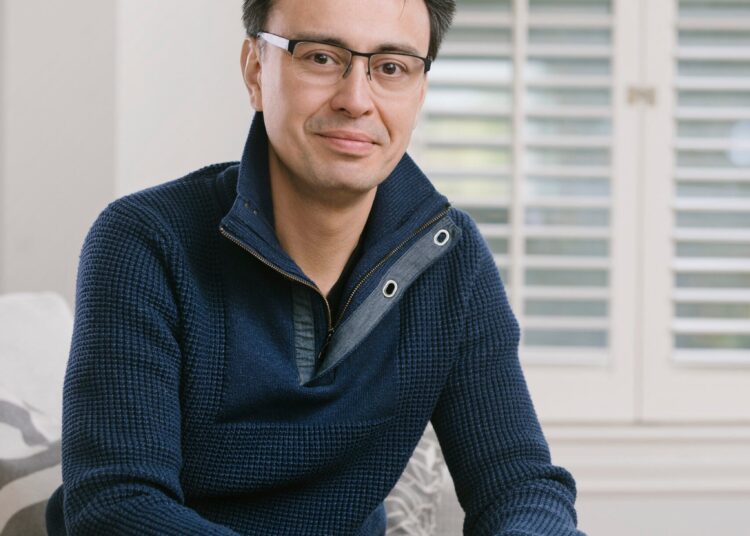 A man with glasses wearing a navy blue sweater sits on a couch, hands clasped, in a room with white shutters and a window in the background.