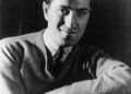 A man with dark hair, wearing a light sweater over a shirt and tie, sits with his arms folded, looking slightly to the side. The photo is in black and white.