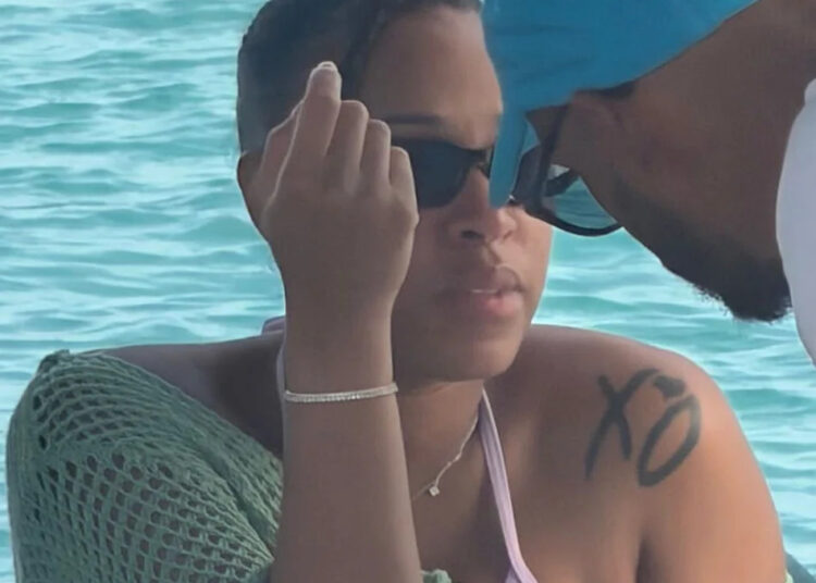 A woman wearing sunglasses and a green cover-up sits by the water. A man in a blue cap leans in toward her.