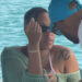 A woman wearing sunglasses and a green cover-up sits by the water. A man in a blue cap leans in toward her.