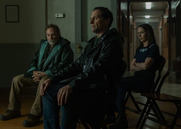 Three people sitting in a dimly lit room, one looking forward, one gazing sideways, and the other with arms crossed.