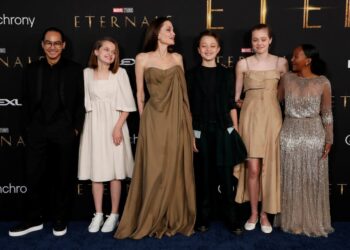 A woman stands in front of a movie backdrop with 2 children on her left and 3 children on her right. All are wearing formal attire.