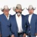Five members of Cautivo Norteño wearing blue blazers and cowboy hats pose together in a studio portrait.