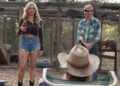 Two people standing outdoors, one holding a bottle, with a person sitting in a hat.