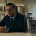 A man in glasses sits at a table counter with a coffee cup near him.