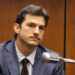 A man in a suit and tie with a mustache sits in a courtroom, looking serious, with a large microphone in front of him and wood-paneled walls in the background.