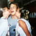 A young man smiles widely as two girls kiss him on the cheeks. All three wear leis and casual clothes, appearing to celebrate in a festive, joyful setting.
