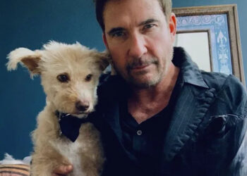 A man in a dark shirt poses indoors next to a small, scruffy tan dog wearing a black bow tie. They are both looking at the camera. There is a blue wall and a framed picture in the background.