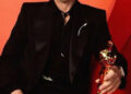 A man in a black suit and glasses smiles while seated on a red chair, holding a gold Oscar statuette against a red background.
