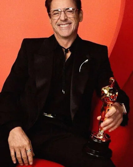 A man in a black suit and glasses smiles while seated on a red chair, holding a gold Oscar statuette against a red background.