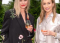 Two women standing outdoors, smiling and holding glasses of rosé wine. One wears a black jacket with floral embroidery, the other a silky light-colored shirt with white bird patterns. Green trees and plants are in the background.