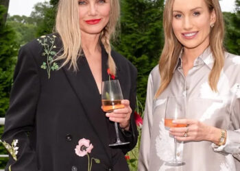 Two women standing outdoors, smiling and holding glasses of rosé wine. One wears a black jacket with floral embroidery, the other a silky light-colored shirt with white bird patterns. Green trees and plants are in the background.