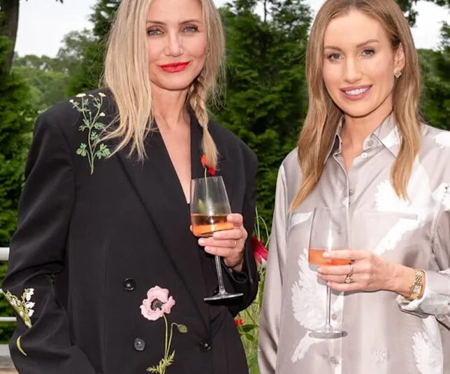 Two women standing outdoors, smiling and holding glasses of rosé wine. One wears a black jacket with floral embroidery, the other a silky light-colored shirt with white bird patterns. Green trees and plants are in the background.