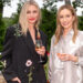 Two women standing outdoors, smiling and holding glasses of rosé wine. One wears a black jacket with floral embroidery, the other a silky light-colored shirt with white bird patterns. Green trees and plants are in the background.