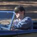 A person wearing a black cap and a blue sweatshirt drives a classic black convertible car with a wooden steering wheel on a sunny day.