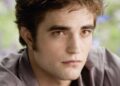 Young person with tousled brown hair and pale skin gazes thoughtfully forward. They wear a gray shirt, and the soft, blurred green and purple background suggests an outdoor setting.