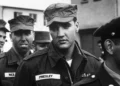A black and white photo of a man in a military uniform with a name tag reading "Presley," standing among other soldiers. The scene appears to be outdoors, possibly during a military event or gathering.