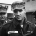 A black and white photo of a man in a military uniform with a name tag reading "Presley," standing among other soldiers. The scene appears to be outdoors, possibly during a military event or gathering.