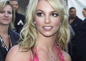 A woman with long blonde hair wearing a pink dress and a cross necklace looks slightly to the right, smiling softly, with people and a white tent blurred in the background.