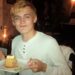 A young person with blond hair, wearing a white shirt, smiles while holding a plate with a slice of cake and a fork, sitting in a dimly lit restaurant.