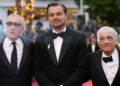 Three men in tuxedos stand together on a red carpet at an event, with blurred lights and trees in the background. They are smiling and appear to be posing for a photo.