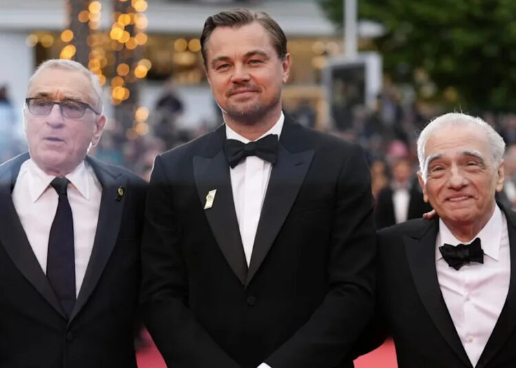 Three men in tuxedos stand together on a red carpet at an event, with blurred lights and trees in the background. They are smiling and appear to be posing for a photo.
