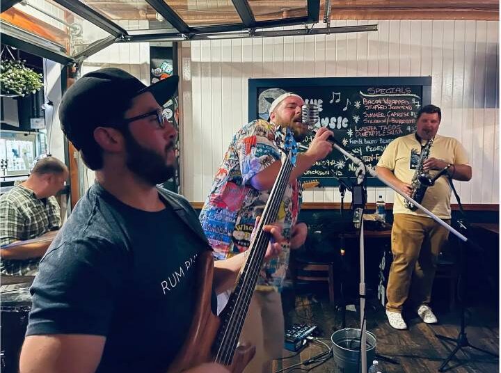 A lively band performing on stage in a casual setting. The bassist is playing while a vocalist sings into a vintage microphone. A saxophonist stands to the right, and another musician is seen in the background. The decor includes a chalkboard menu with daily specials.