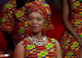 Anna Mudeka, wearing a dress and headdress with a yellow, green, blue and red pattern and a shell necklace, smiles towards a camera while sat down, with other members of a choir wearing similar clothing can be seen behind her.