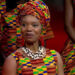 Anna Mudeka, wearing a dress and headdress with a yellow, green, blue and red pattern and a shell necklace, smiles towards a camera while sat down, with other members of a choir wearing similar clothing can be seen behind her.