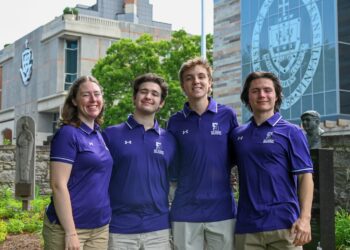 Four students in matching purple polo shirts pose outdoors in front of campus buildings and a large university seal on a glass facade.