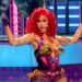Cardi B performing on stage with bright red hair, wearing a colorful, intricate outfit. She's energetically dancing with backup dancers.