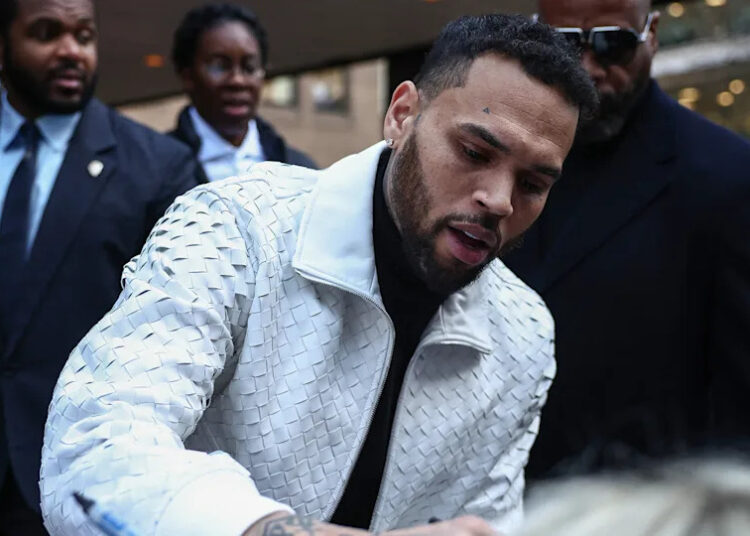 Chris Brown, in a textured white jacket, signing an autograph, surrounded by people in formal attire outdoors.