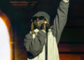 Lil Wayne performing on stage, wearing sunglasses, a gray hat, and a jacket, with one hand raised and holding a microphone.