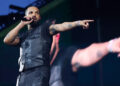 Drake performing on stage, wearing a black vest, holding a microphone, and pointing towards the audience.