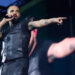Drake performing on stage, wearing a black vest, holding a microphone, and pointing towards the audience.