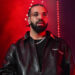 Drake with a beard and glasses performs on stage, wearing a black leather jacket, with red lighting in the background.