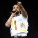 Drake performing on stage, wearing a white vest with graphics, holding a microphone, and pointing towards the audience.