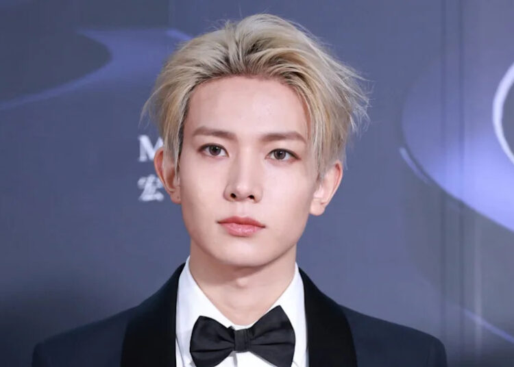 K-pop star Heeseung with blonde hair in a tuxedo and bow tie at a formal event, standing against a dark background.