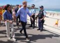 A man and a woman walking near a beach, as multiple people, including police, look on.