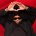 Druski makes a triangle shape with his hands against a red background, parodying Jay-Z.