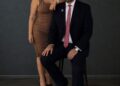 A woman and man in formal attire pose confidently against a dark background.