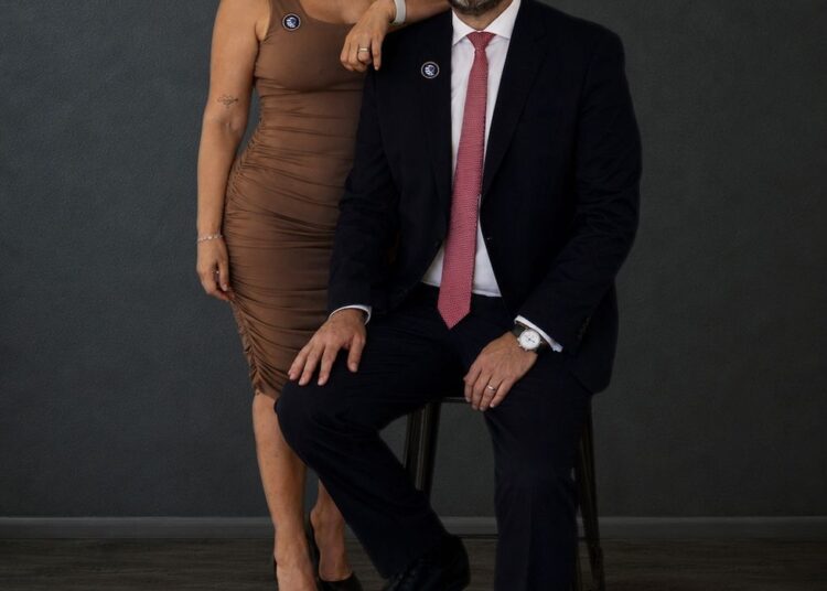 A woman and man in formal attire pose confidently against a dark background.